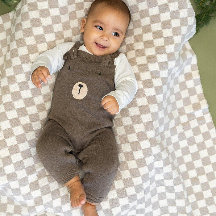 Baby wearing a brown romper with a checkered blanket and greenery in the background