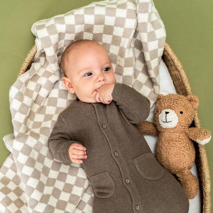 A baby wearing a brown knit footed one-piece romper lies in a woven bassinet with a beige-and-white check blanket and a brown teddy bear, against an olive green background.