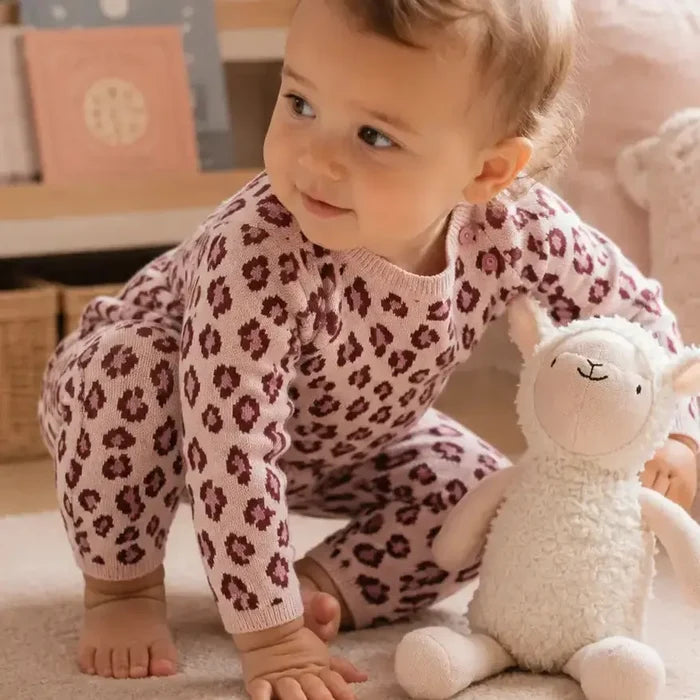 A baby crawling on a carpeted floor next to a plush white stuffed animal, wearing a pink and burgundy leopard print knit jumpsuit.