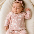 A sleeping newborn baby wearing a pink long-sleeve wrap-style sleeper with white cloud and star prints, matching footed pants, and a pink bow headband, lying on a cream baby lounger.