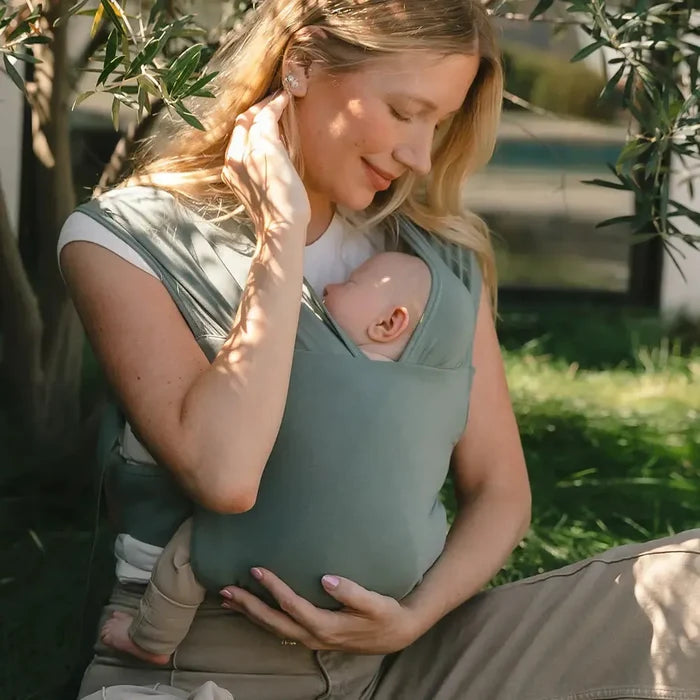 Woman sitting outdoors in a grassy area, holding a baby in a green carrier.