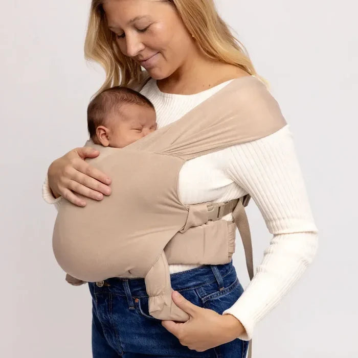 Woman holding a baby in a beige baby carrier against a plain background