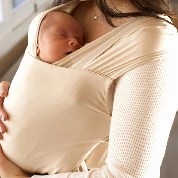 Woman holding a baby wrapped in a beige sling indoors.