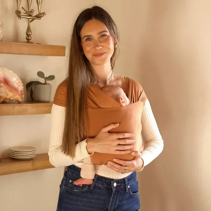 Woman carrying a baby in a brown sling against a beige wall with wooden shelves.