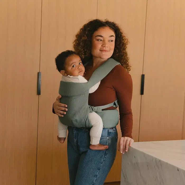 Woman holding a baby in a carrier in a room with wooden walls and a concrete floor.