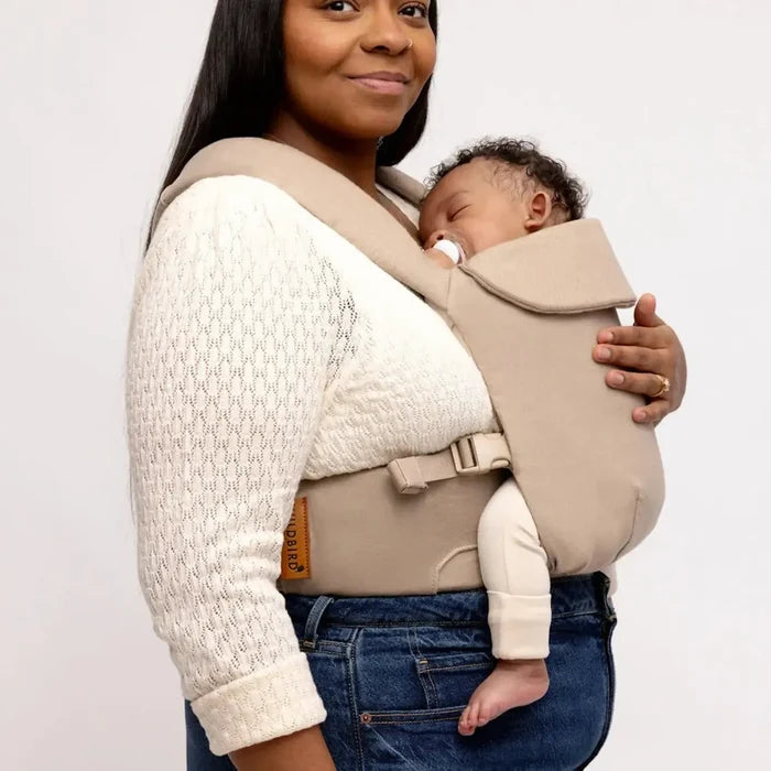 Woman holding a baby in a beige baby carrier against a plain background