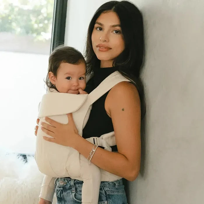 Woman holding a baby in a carrier against a light-colored wall.