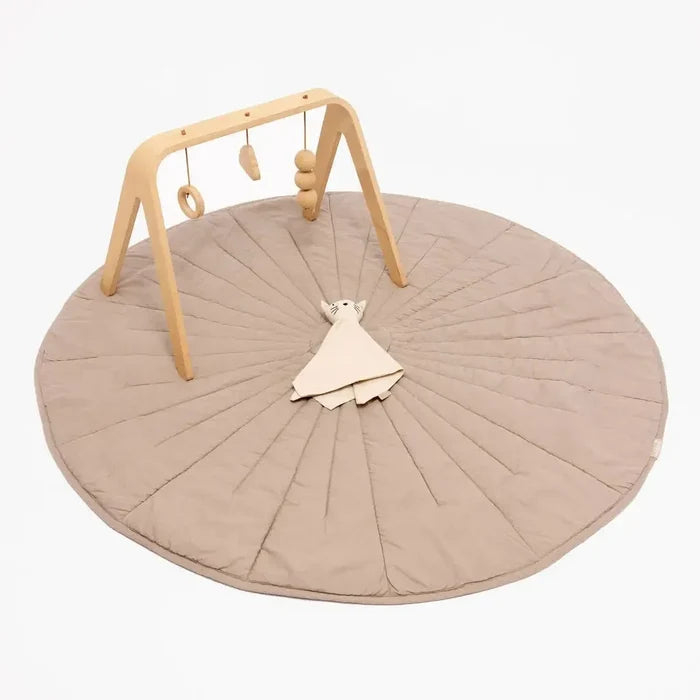 A baby activity gym set featuring a large round quilted play mat in a warm taupe-beige color, a natural wood arch with hanging wooden toys, and a matching beige plush comforter, all photographed on a clean white background.