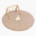 A baby activity gym set featuring a large round quilted play mat in a warm taupe-beige color, a natural wood arch with hanging wooden toys, and a matching beige plush comforter, all photographed on a clean white background.