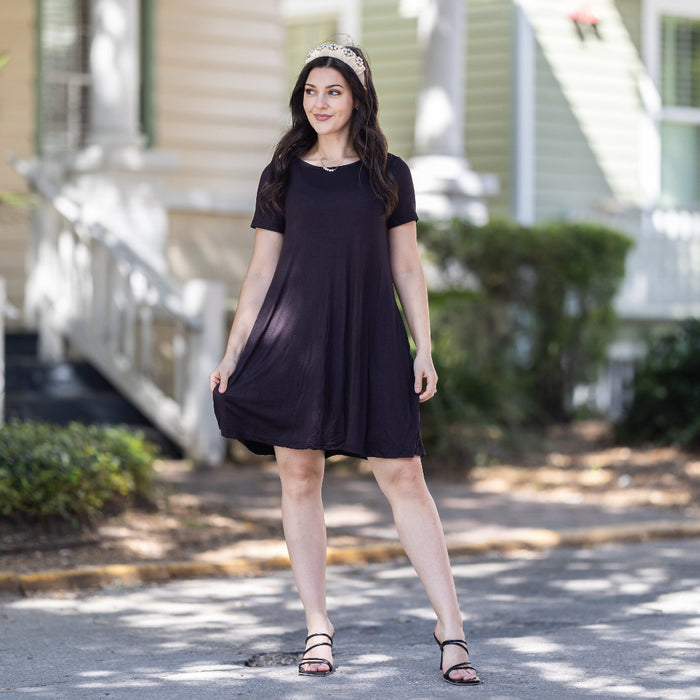 A woman standing on a street wearing a black short sleeve swing dress with pockets, paired with white sandals.